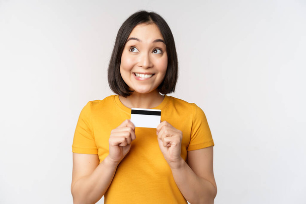 Retrato de bela menina coreana segurando cartão de crédito, recomendando serviço bancário, de pé em camiseta amarela sobre fundo branco. - Foto, Imagem