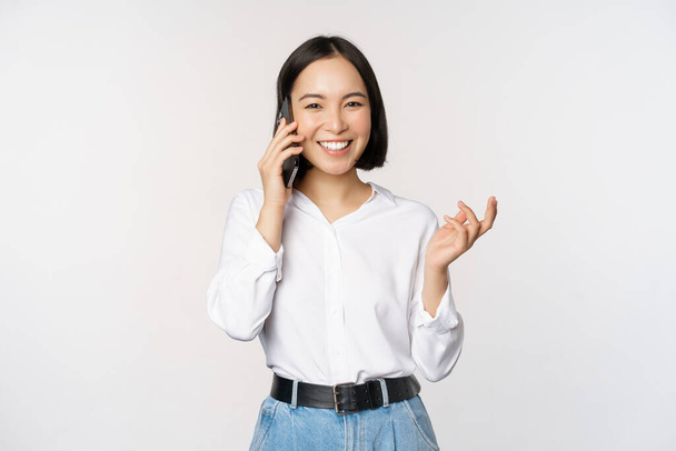 Sonriente mujer asiática feliz hablando en el teléfono inteligente con el cliente, vendedora de guardia, sosteniendo el teléfono móvil y el gesto, de pie sobre fondo blanco. - Foto, imagen