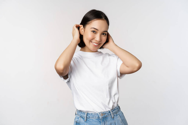 Retrato de linda, hermosa mujer asiática tocando su nuevo corte de pelo corto, mostrando el peinado, sonriendo feliz en la cámara, de pie sobre fondo blanco. - Foto, Imagen