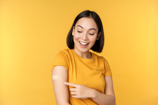 Vacunación y concepto de pandemia covid-19. Feliz y saludable chica asiática apuntando a su hombro con curita después de vacunar de coronavirus, fondo amarillo. - Foto, imagen