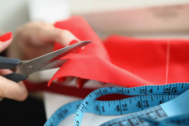 Female hands with manicure cut red fabric with scissors close-up. There is a cloth and a measuring tape on the table - 写真・画像