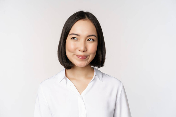 Image d'une femme asiatique souriante planifiant, pensant à smth, rêvant, debout sur fond blanc avec un visage suffisant. Espace de copie - Photo, image