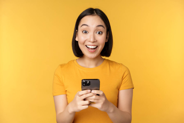 Šťastná asijská dívka se usmívá, stojí s černým mobilním telefonem, stojí na žlutém pozadí. Kopírovat prostor - Fotografie, Obrázek
