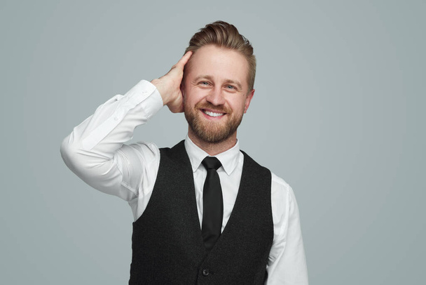Positive bearded businessman in white shirt and black vest looking at camera with toothy smile and touching blond hair against gray background - Foto, imagen