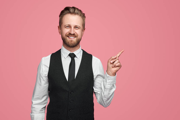 Positive bearded male entrepreneur in white shirt and black vest looking at camera with smile and pointing away against pink background - Foto, imagen
