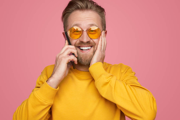 Excited male hipster receiving good news and speaking on mobile phone while touching cheek and looking at camera on pink background - Foto, Imagen