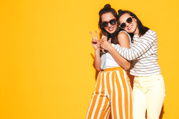 Two young beautiful smiling blond hipster female in trendy summer clothes. Sexy carefree women posing near yellow wall in studio. Positive models having fun. Cheerful and happy. In sunglasses - Foto, imagen
