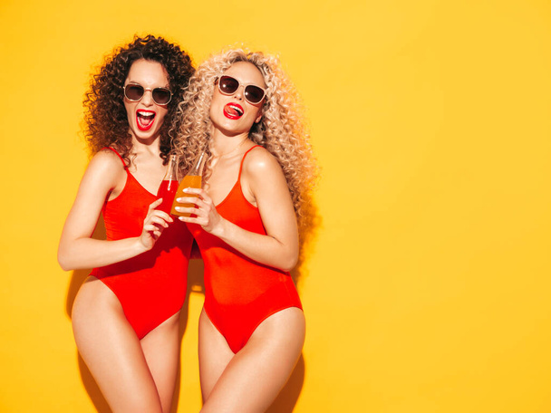 Two beautiful sexy smiling hipster women in red summer swimwear bathing suits.Trendy models with afro curls hairstyle having fun in studio.Hot female isolated on yellow.Drinking lemonade from bottle - Fotografie, Obrázek