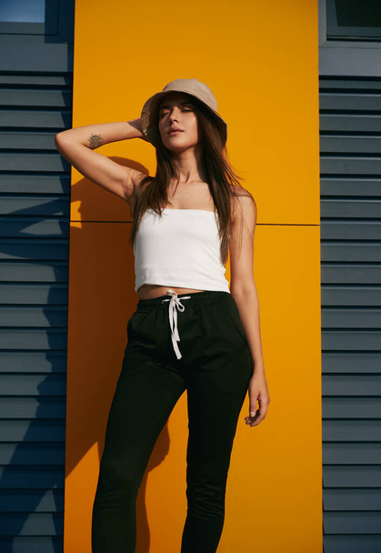 Trendy young female in casual clothes touching hat and enjoying sunny day while standing near wall of modern building on street - Foto, afbeelding