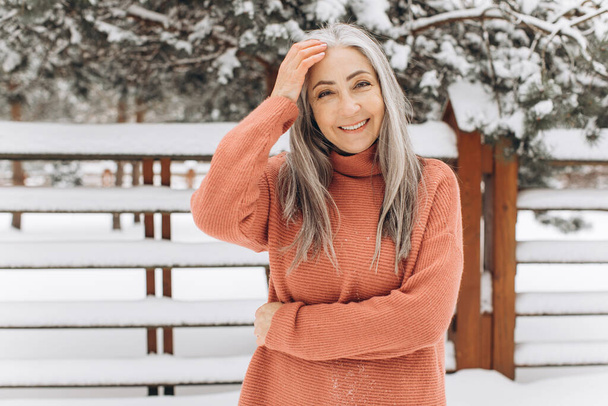 Femme âgée avec les cheveux gris dans un pull tricoté, souriant sincèrement sur un fond d'hiver. - Photo, image