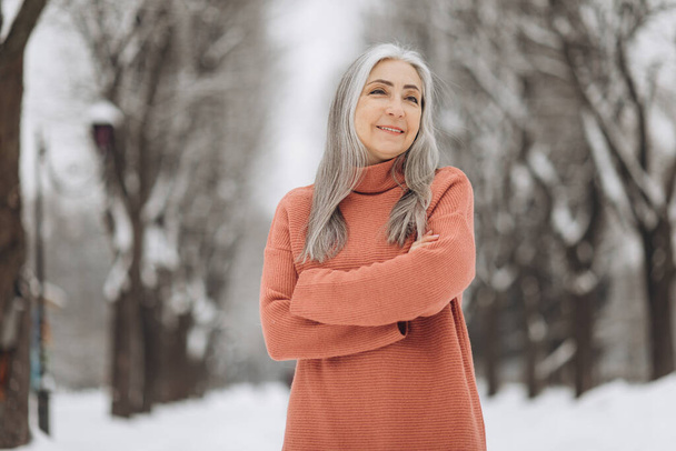 Portrét starší ženy s šedými vlasy v pleteném svetru s úsměvem na pozadí sněžného počasí v zimě - Fotografie, Obrázek