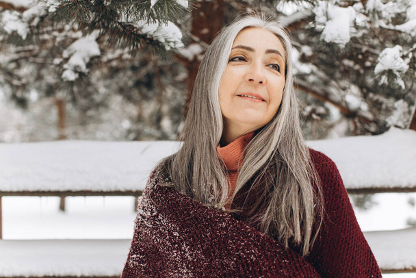 Femme âgée avec des cheveux gris dans un pull tricoté et une écharpe, souriant sincèrement sur un fond d'hiver. - Photo, image