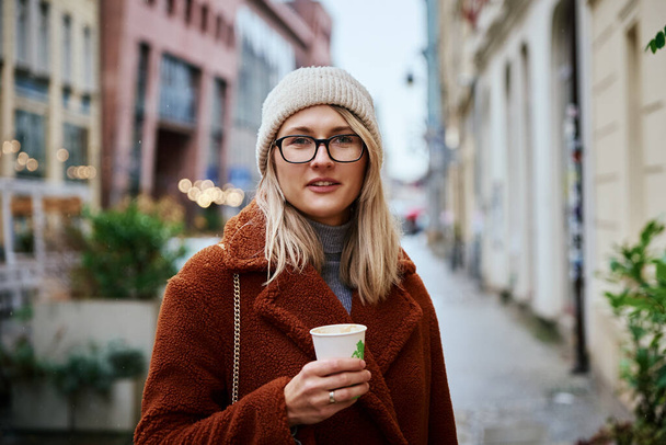 Jonge vrouw in bril draagt jas lopen op straat met een kop koffie afhaalmaaltijden - Foto, afbeelding