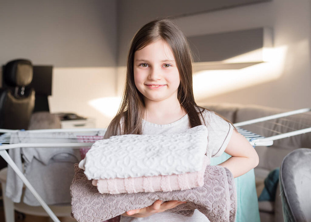 The girl is holding a clean folded towel in her hands. Concept help children around the house. Little girl helps her mother to fold laundry - Photo, Image