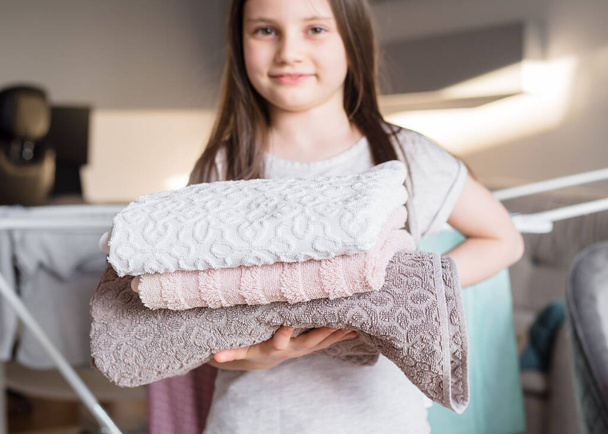 La chica sostiene una toalla doblada limpia en sus manos. Concepto de ayudar a los niños de la casa. Niña ayuda a su madre a doblar la ropa - Foto, imagen