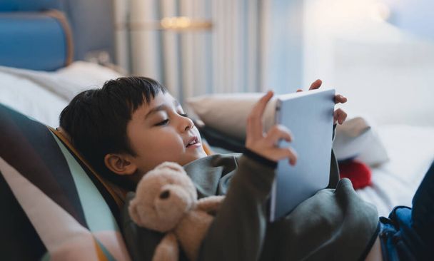 Happy Kid makaa sängyssä tilalla tabletti katsomassa sarjakuva ja jutella ystävien kanssa digitaalisessa pad, Söpö nuori poika pelaa pelejä verkossa internetissä, Lapsi rentouttava kotona perheen kanssa - Valokuva, kuva