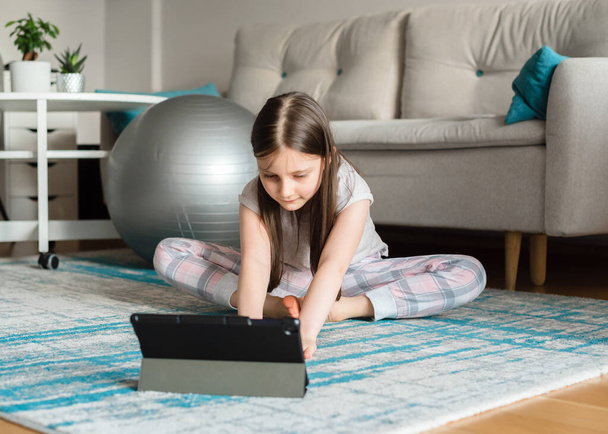 Menina treina on-line em um tablet para alongamento com um fitball. Atividades esportivas durante a pandemia do coronovírus. Quarentena. Estudo online - Foto, Imagem