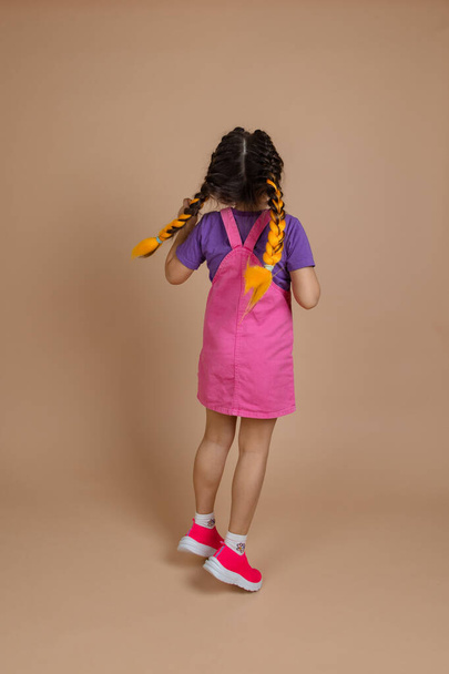 Back of energetic, little female kid with two kanekalon pigtails of yellow color, jumping in pink jumpsuit, purple t-shirt and pink sneakers on beige background - Valokuva, kuva