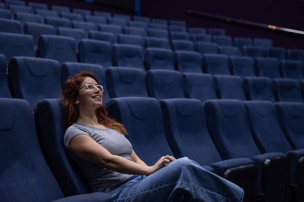 Caucasian red-haired woman sits on the front row in a cinema in an empty hall. The girl is watching a movie alone.  - Fotografie, Obrázek