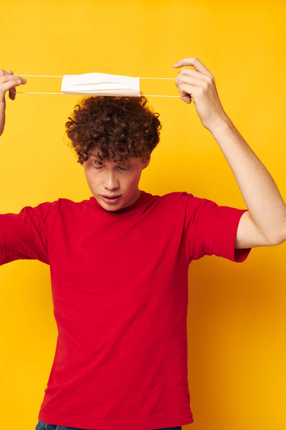 portrait of a young curly man wearing a red t-shirt medical mask on the face posing yellow background unaltered - Foto, Bild