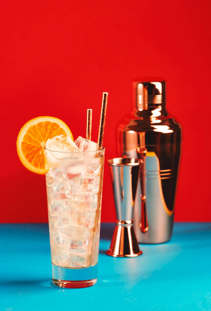 Tom Collins alcoholic cocktail with dry gin, syrup, lemon juice, soda, orange and ice. Defiant red blue background, bar tools, copy space - Фото, изображение