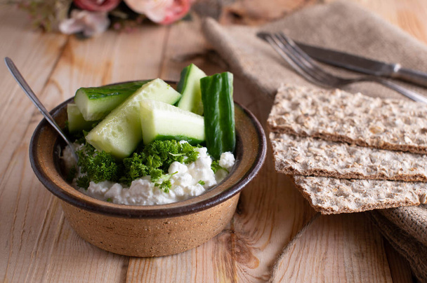 Homemade healthy meal or snack with cottage cheese, chopped herbs and cucumber. Served in a stoneware bowl with crispbread on wooden table. Ready to eat - Photo, Image