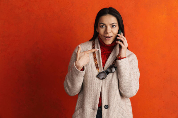 Young hispanic woman pointing finger aside while talking on cellphone isolated over orange background - Foto, imagen
