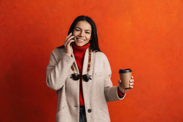 Smiling hispanic woman drinking coffee while talking on mobile phone isolated over orange wall - 写真・画像