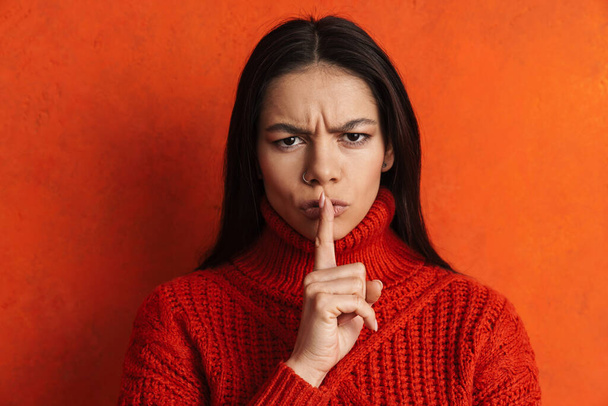 Displeased hispanic woman frowning and showing silence gesture isolated over orange wall - 写真・画像