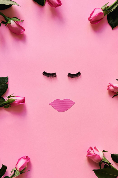 Falsas pestañas y labios de papel sobre fondo rosa con flores de color rosa. Una cama plana femenina. Concepto de belleza - Foto, imagen