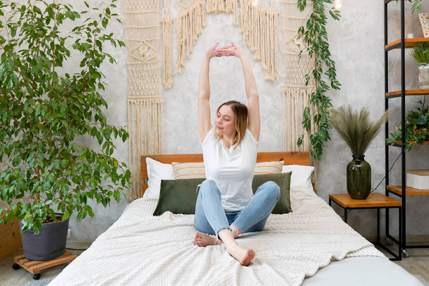 Photo of young happy woman stretching her arms and smiling while sitting on bed - Foto, afbeelding