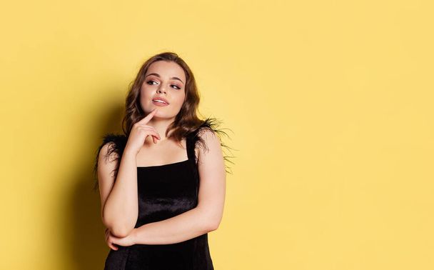 Studio shot of young pretty girl with long curly hair wearing black evening dress isolated on yellow background. Concept of emotions, beauty, fashion - Valokuva, kuva