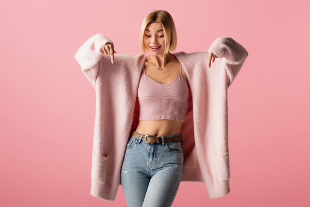 happy young woman in soft cardigan and jeans pointing down with fingers isolated on pink - Foto, Imagem