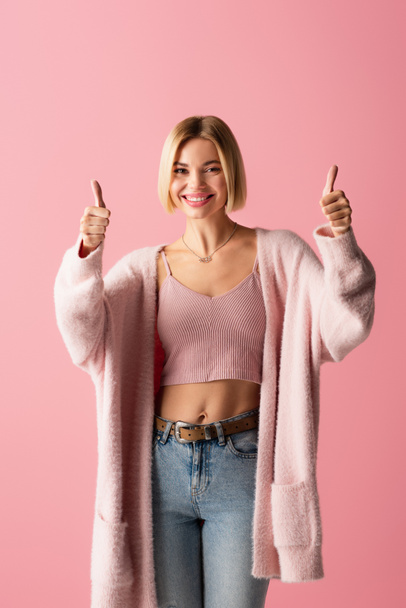 happy young woman in soft cardigan showing thumbs up isolated on pink  - Фото, зображення