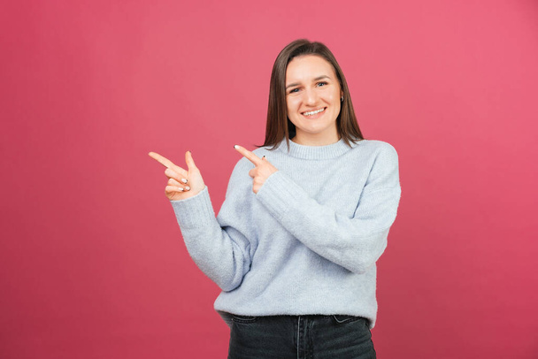 Joven mujer sonriente señala a un lado con ambos dedos índice sobre fondo rosa en un estudio. - Foto, imagen