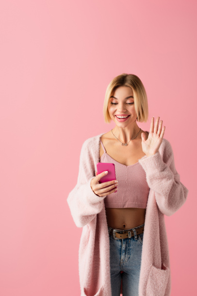 heureuse jeune femme en doux cardigan agitant la main lors d'un appel vidéo isolé sur rose  - Photo, image