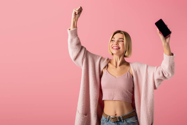 happy young woman in soft cardigan holding smartphone with blank screen isolated on pink  - Zdjęcie, obraz