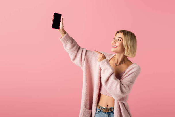 glückliche junge Frau in weicher Strickjacke zeigt auf Smartphone mit leerem Bildschirm isoliert auf rosa  - Foto, Bild