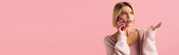 displeased young woman in soft cardigan talking on smartphone isolated on pink, banner - Photo, Image