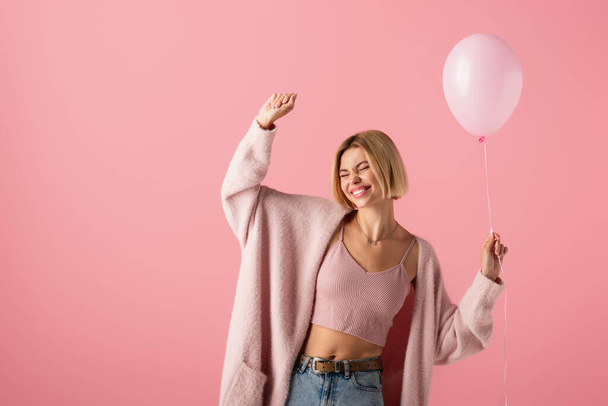 joyful young woman in cardigan holding balloon isolated on pink - Fotó, kép