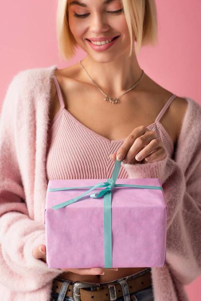cheerful young woman in cardigan pulling blue ribbon on wrapped gift box isolated on pink - Foto, afbeelding