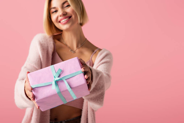 blurred and cheerful young woman in cardigan holding wrapped gift box isolated on pink - Φωτογραφία, εικόνα