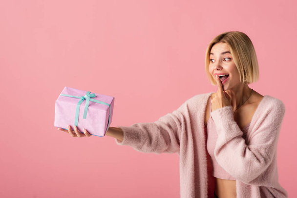 excited young woman in cardigan covering mouth with finger while holding wrapped gift box isolated on pink - Foto, Imagem