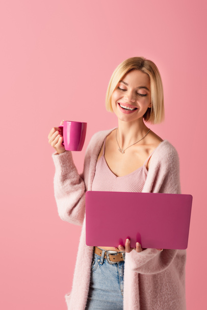 χαρούμενη γυναίκα κρατώντας κούπα με καφέ και φορητό υπολογιστή απομονωμένο σε ροζ  - Φωτογραφία, εικόνα