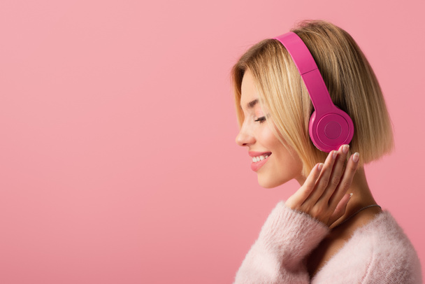 side view of cheerful blonde woman listening music in wireless headphones isolated on pink - Fotografie, Obrázek