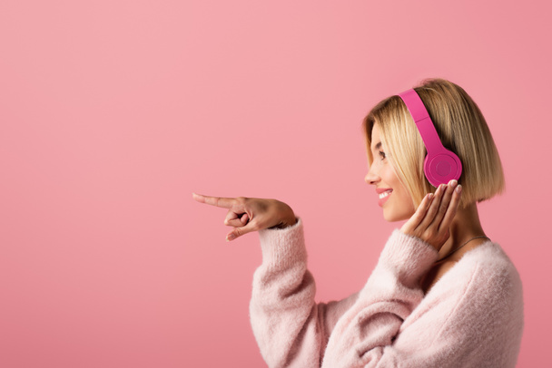 oldalnézetben vidám szőke nő hallgat zenét vezeték nélküli fejhallgató és ujjal elszigetelt rózsaszín - Fotó, kép
