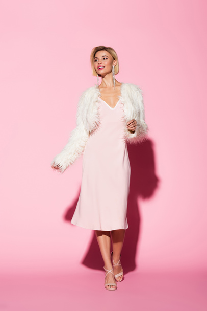 full length of stylish young woman in slip dress and faux fur jacket smiling on pink  - Фото, изображение