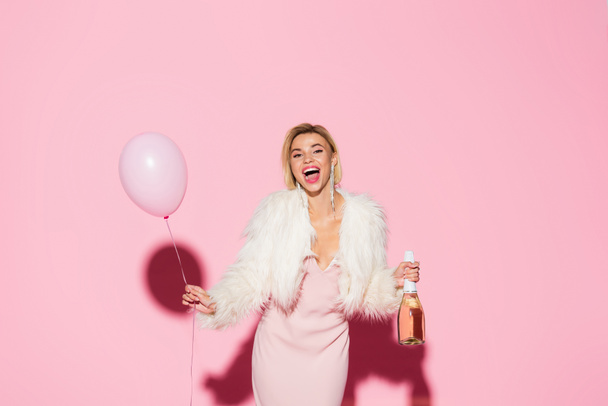 excited woman in slip dress and faux fur jacket holding balloon and bottle of champagne on pink  - Fotografie, Obrázek