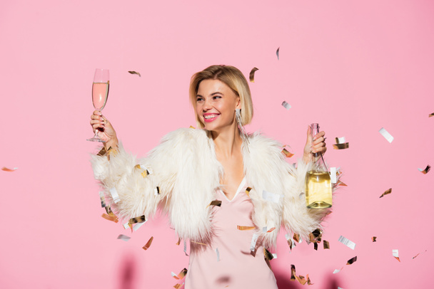 cheerful woman in slip dress and faux fur jacket holding bottle of champagne and glass near falling confetti on pink  - Fotoğraf, Görsel