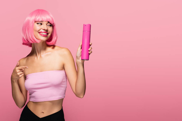 femme gaie avec des cheveux colorés pointant vers la bouteille avec vaporisateur isolé sur rose  - Photo, image
